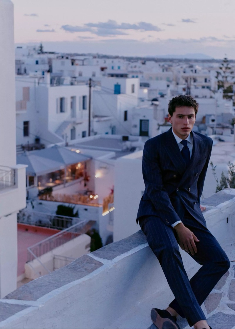 Clément perches on a low rooftop wall above a Greek town at dusk in a navy pinstripe double-breasted suit with a navy tie and suede loafers for Mango Selection's spring-summer 2026 collection