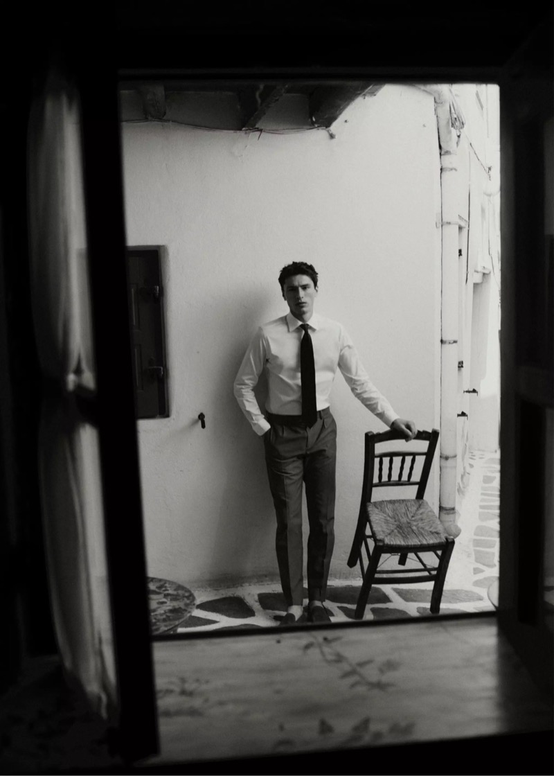 Black and white image framed through a doorway of Clément leaning on a chair in a white shirt, dark tie, and gray trousers for Mango Selection's spring-summer 2026 collection