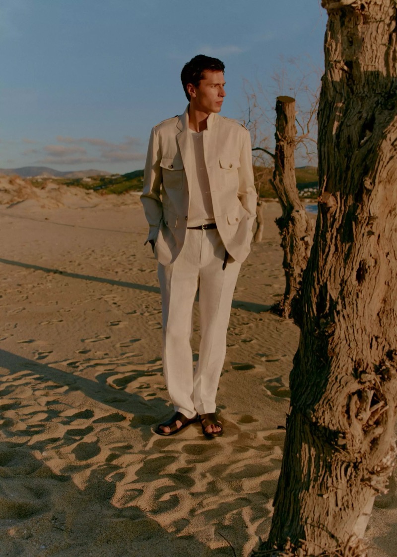 Clément stands on a beach beside a bare tree in a cream safari-style field jacket over a cream knit and white linen trousers with dark sandals for Mango Selection's spring-summer 2026 collection