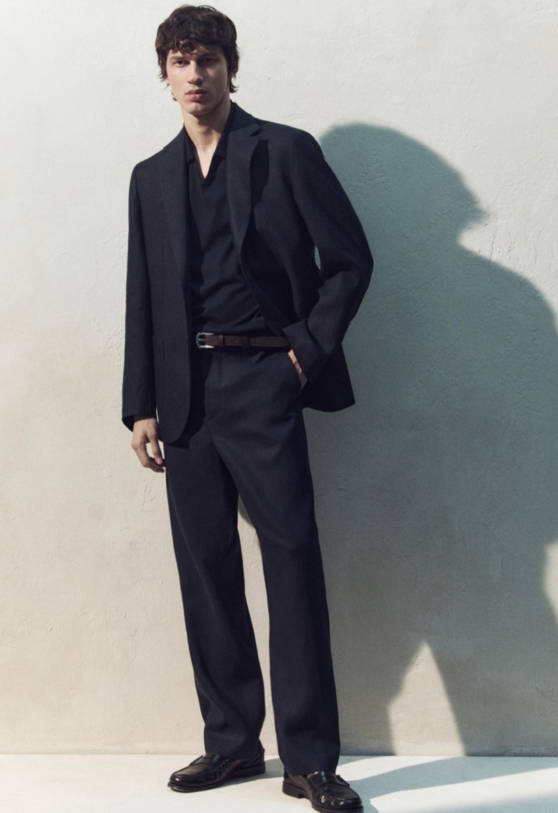Kaplan wears a navy suit with a matching open-collar shirt and brown leather belt