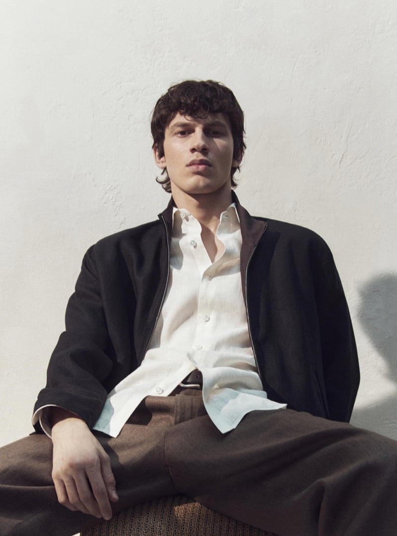 Kaplan wears a black zip jacket over a white shirt with brown trousers
