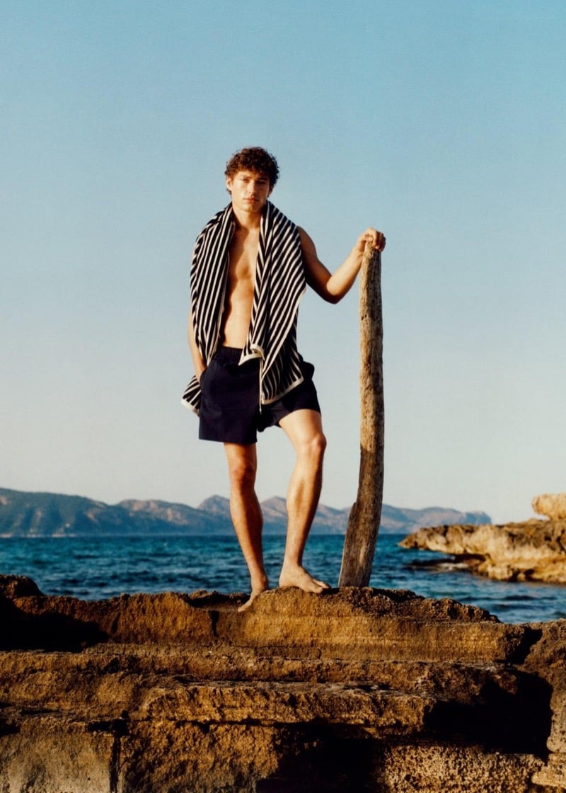 Valentin stands on the rocky coast with a striped towel over his shoulders and navy Massimo Dutti swim shorts