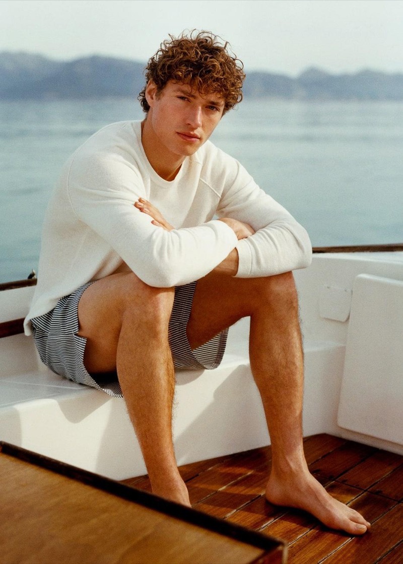 Valentin sits on the deck of a wooden sailboat in a white crew neck sweater and striped Massimo Dutti swim shorts