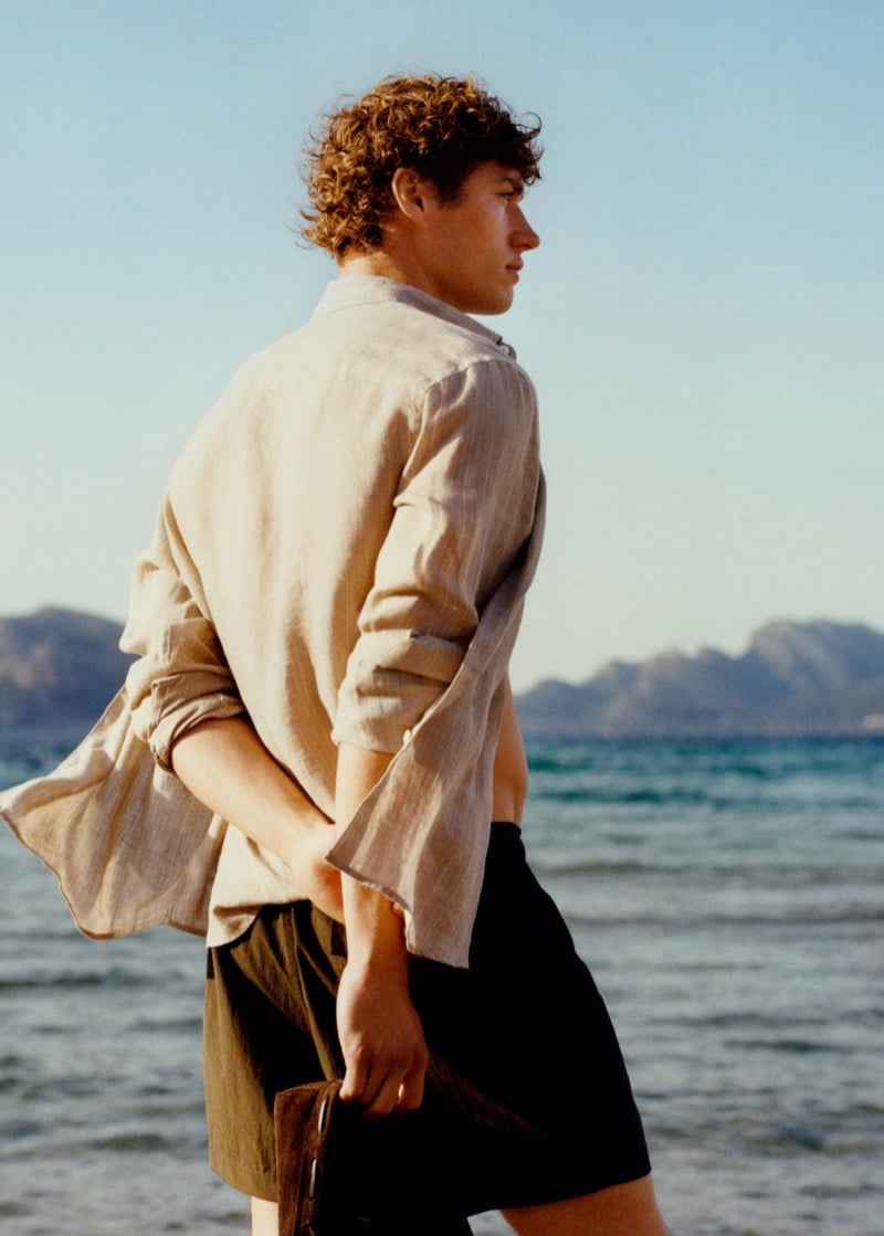 Valentin walks along the coast seen from behind in a linen shirt and dark Massimo Dutti swim shorts carrying a leather pouch