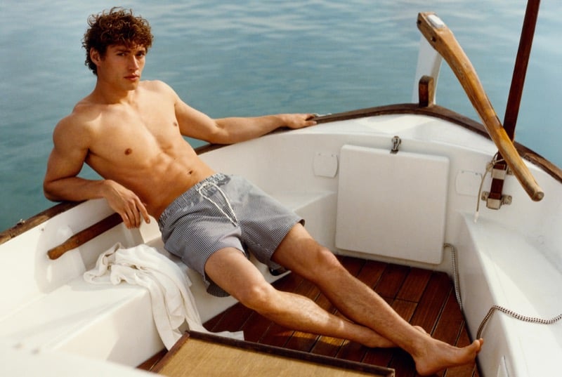 Valentin reclines shirtless in a wooden sailboat wearing striped Massimo Dutti swim shorts