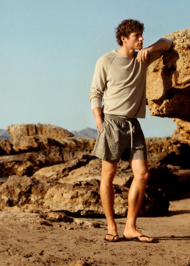 Valentin leans against a rock in a crew neck sweater and textured gray Massimo Dutti swim shorts with flip-flops