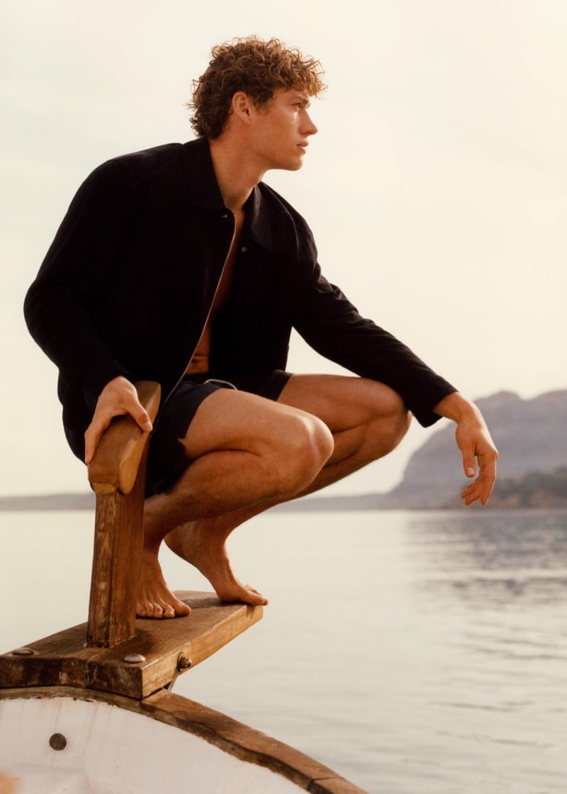 Valentin crouches on the prow of a wooden sailboat in an open black overshirt and dark Massimo Dutti swim shorts