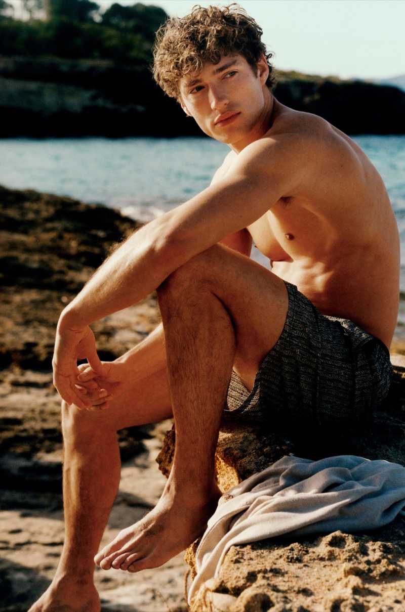 Valentin sits shirtless on the rocky coast at golden hour in dark textured Massimo Dutti swim shorts