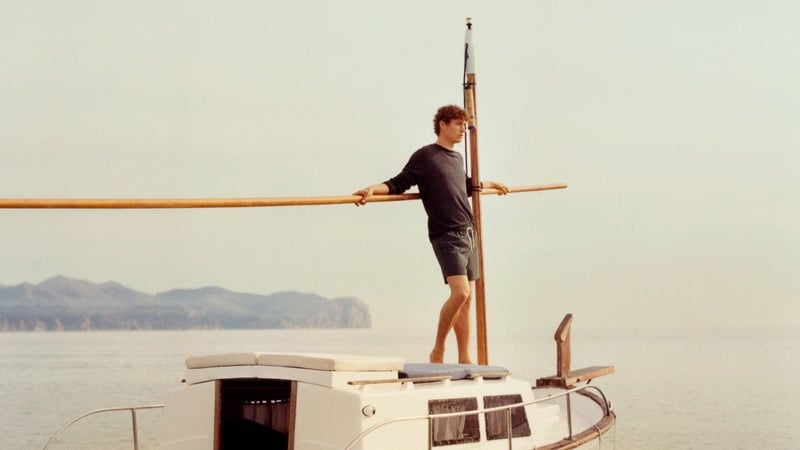 Valentin stands at the mast of a wooden sailboat in a navy sweatshirt and dark Massimo Dutti shorts