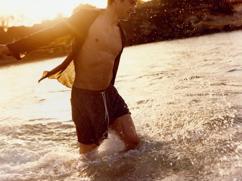 Valentin wades through the surf at sunset with an open shirt billowing behind him in dark printed Massimo Dutti swim shorts