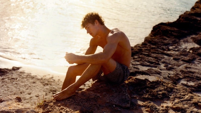 Valentin sits on the rocky coast at sunset in dark textured Massimo Dutti swim shorts with golden backlight