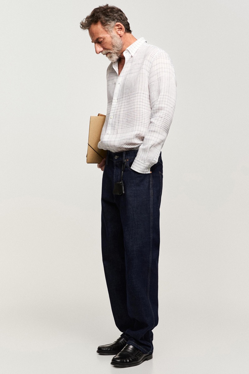 Alain wears a white grid-check shirt with dark jeans in profile