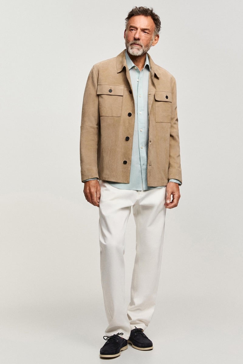 Alain wears a tan suede overshirt over a light blue shirt with white trousers