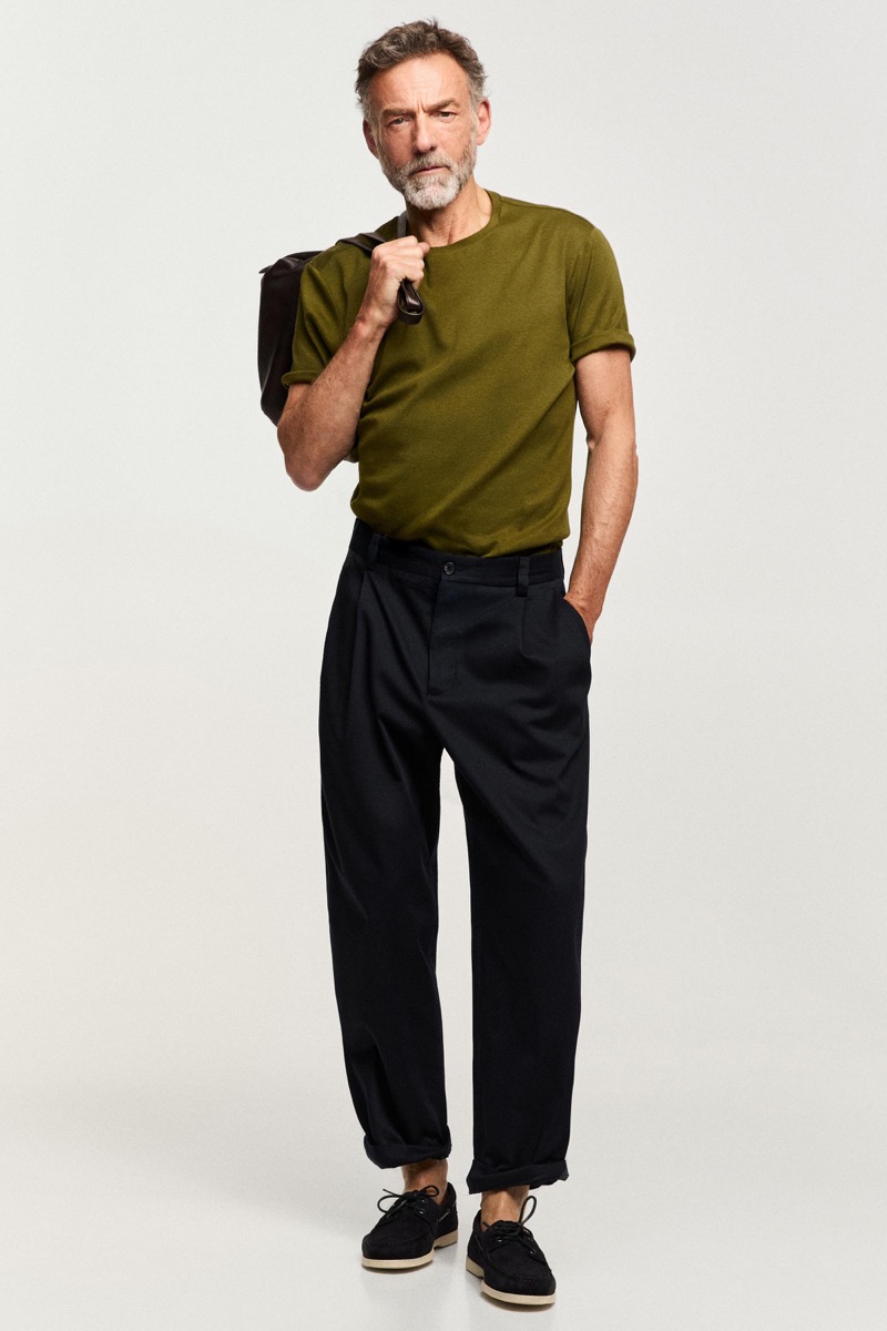 Alain wears an olive T-shirt with dark pleated trousers and a bag