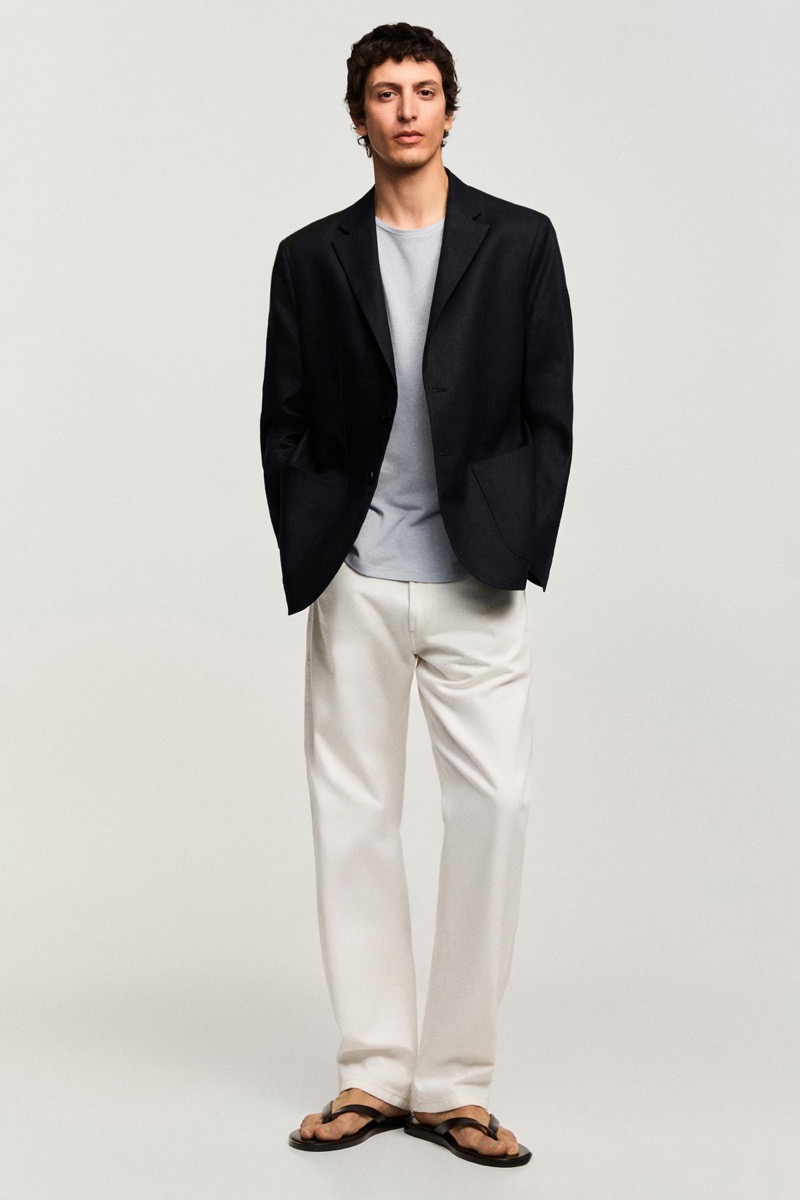 Takfarines wears a black softened blazer over a gray tee with white trousers and sandals