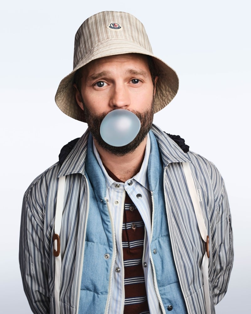 Dornan blows a blue bubble gum bubble in a beige pinstripe Moncler bucket hat and layered striped shirt jacket over a blue down gilet