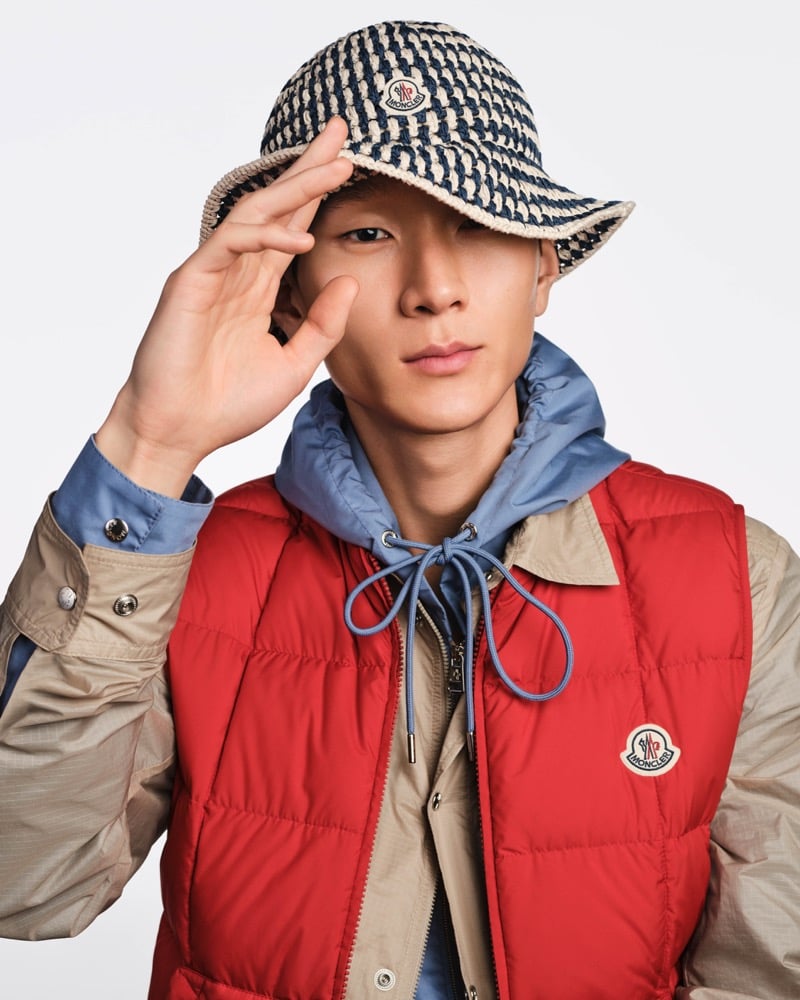 Jun Yeong holds the brim of a navy and cream woven Moncler bucket hat over a red down gilet and blue hooded windbreaker