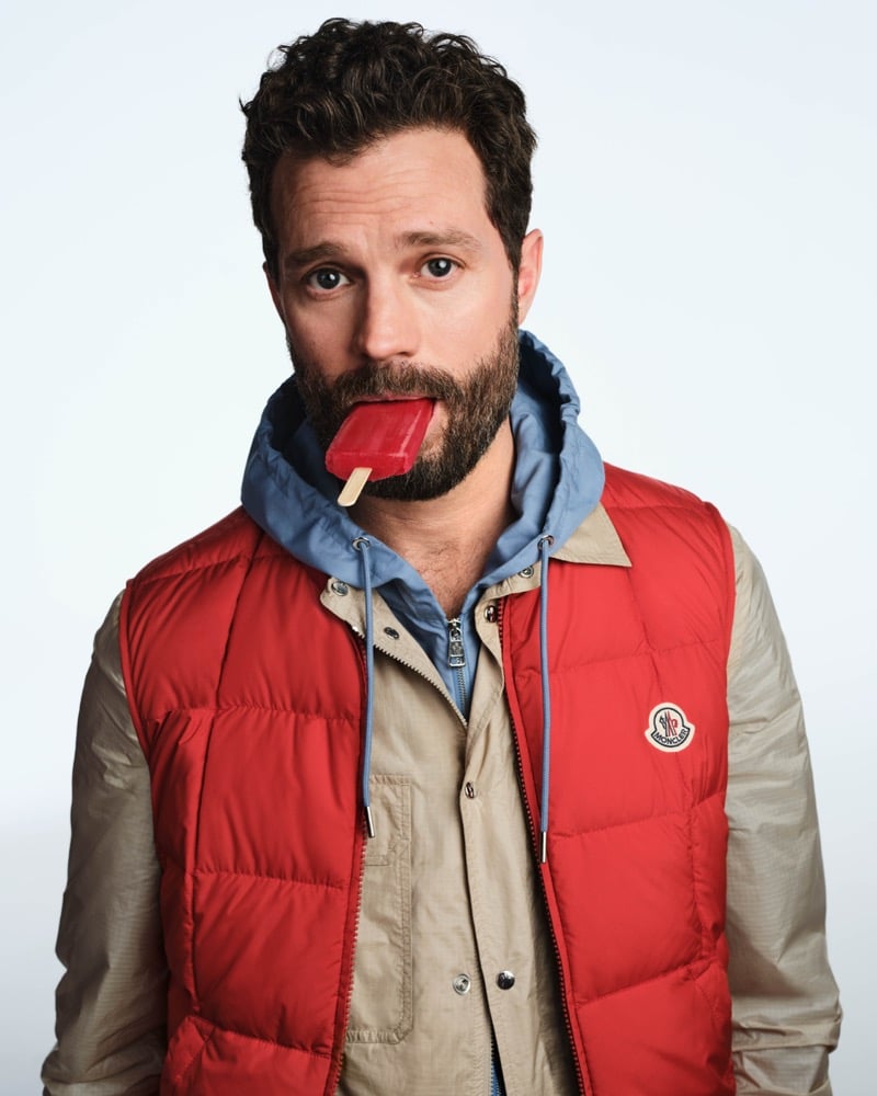 Jamie bites a red popsicle in a red Moncler down gilet layered over beige and blue jackets