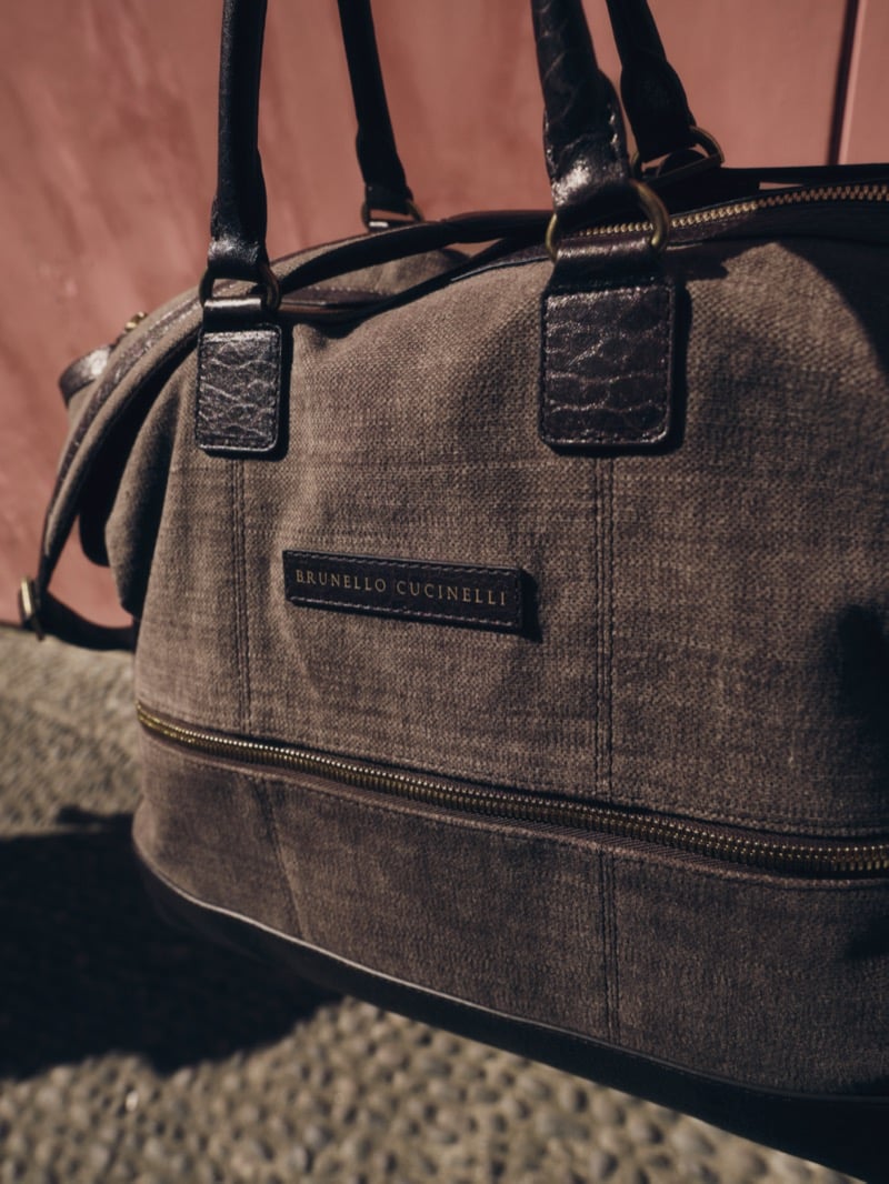 Brunello Cucinelli leather-trimmed woven weekend bag with brass hardware against a terracotta wall