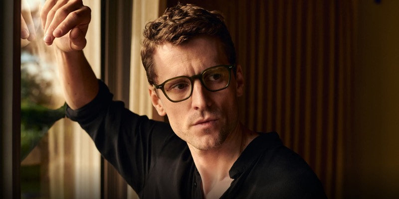 George wears Oliver Peoples Arllett olive acetate optical glasses in a black shirt beside wood paneling