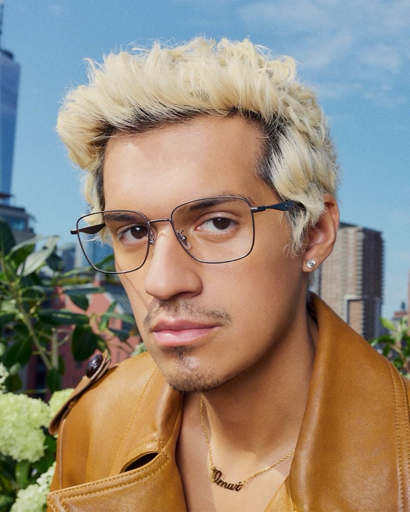Omar Apollo wears Coach C Hardware square optical eyeglasses in gunmetal with a tan leather jacket and gold nameplate necklace, photographed on a rooftop