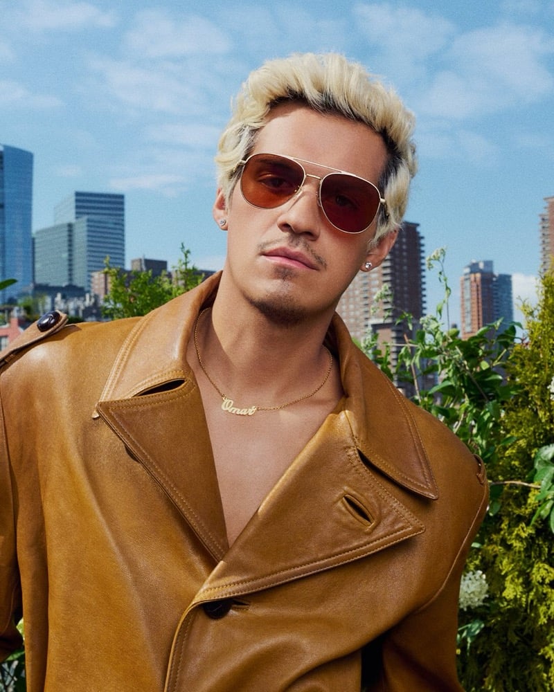 Apollo wears Coach C Hardware Metal Pilot sunglasses and a thin metal frame, paired with a tan leather jacket on a New York rooftop