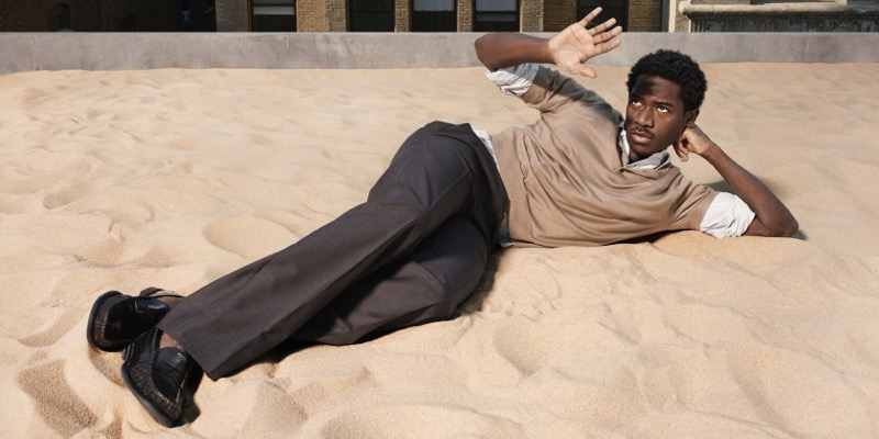 Idris reclines on sand atop a city rooftop in charcoal wool trousers, a tan open-knit cotton polo over a white shirt, and black leather loafers for Prada's Days of Summer 2026 campaign