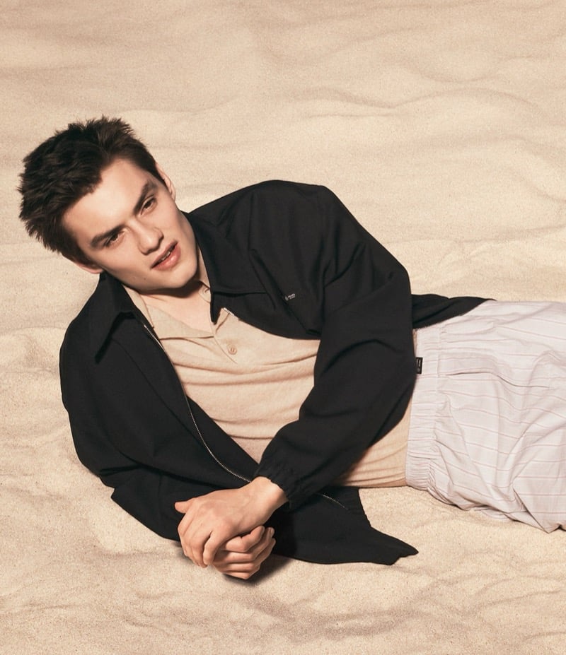 Partridge lies on rooftop sand in a black Prada zip-up jacket over a tan polo and pinstriped lounge shorts for Prada's Days of Summer 2026 campaign