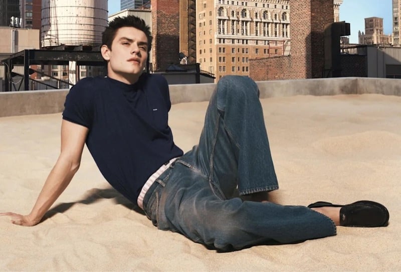 Partridge reclines on rooftop sand in a navy crew neck T-shirt and loose faded jeans with black leather loafers, skyscrapers behind him, for Prada's Days of Summer 2026 campaign