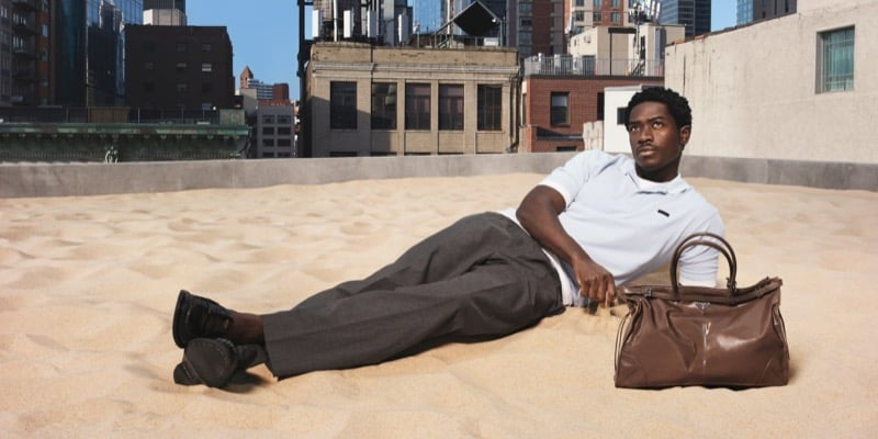 Idris lies back on rooftop sand in a white Prada knit polo and charcoal wool trousers with black leather loafers beside a brown leather duffel bag, the city skyline behind him, for Prada's Days of Summer 2026 campaign