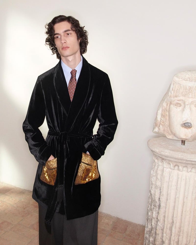 Sombr wears a black velvet Valentino robe with gold sequin pockets, a blue shirt, and a patterned brown tie beside an ancient carved stone bust on a pedestal for the pre-fall 2026 campaign