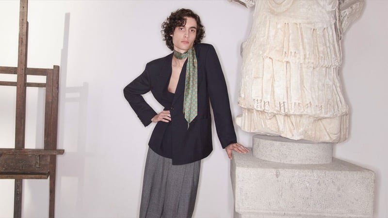 Sombr wears an open Valentino blazer over a bare chest with a green patterned silk scarf around the neck and gray wool trousers beside a draped classical torso sculpture for the pre-fall 2026 campaign