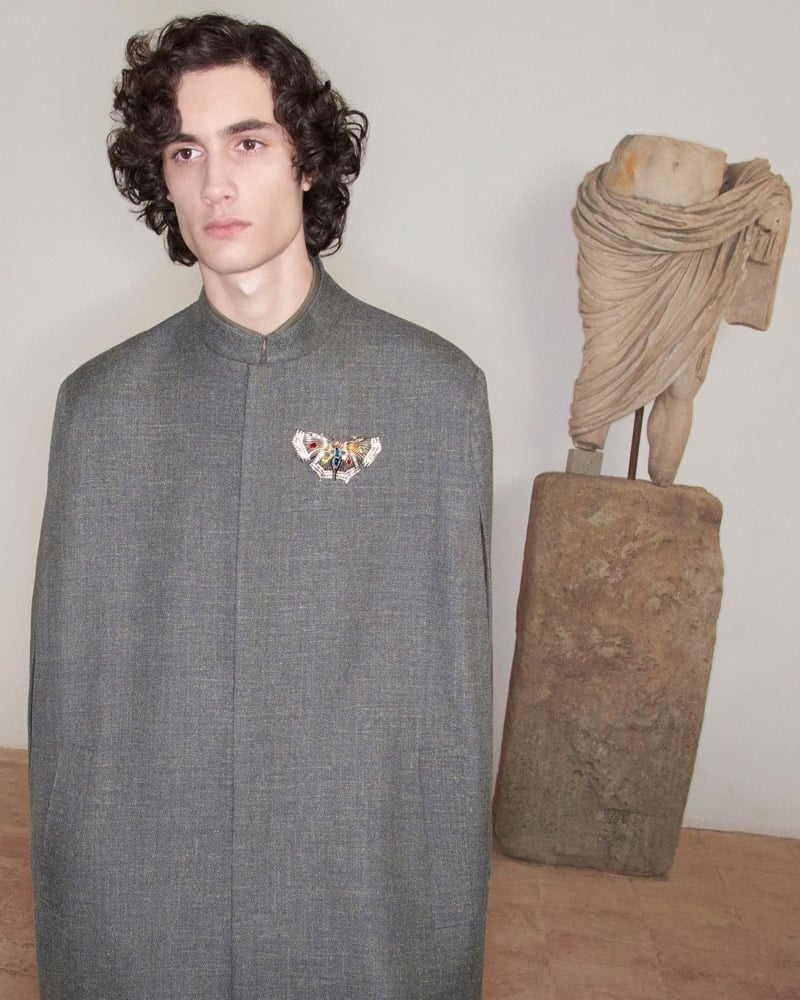 Sombr wears a gray wool Valentino cape with a beaded butterfly brooch beside a fragment of a draped stone torso for the pre-fall 2026 campaign