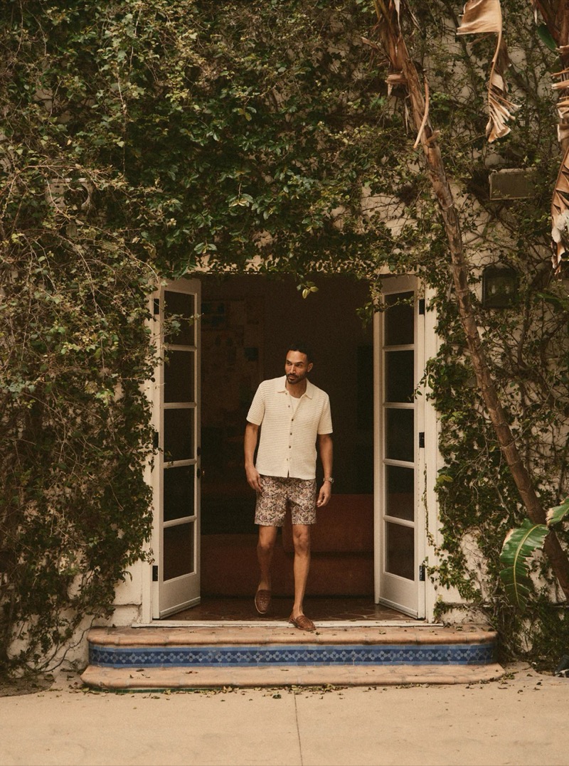 A man in a cream geometric-weave short-sleeve shirt and printed shorts stands in an ivy-covered doorway for Taylor Stitch summer 2026