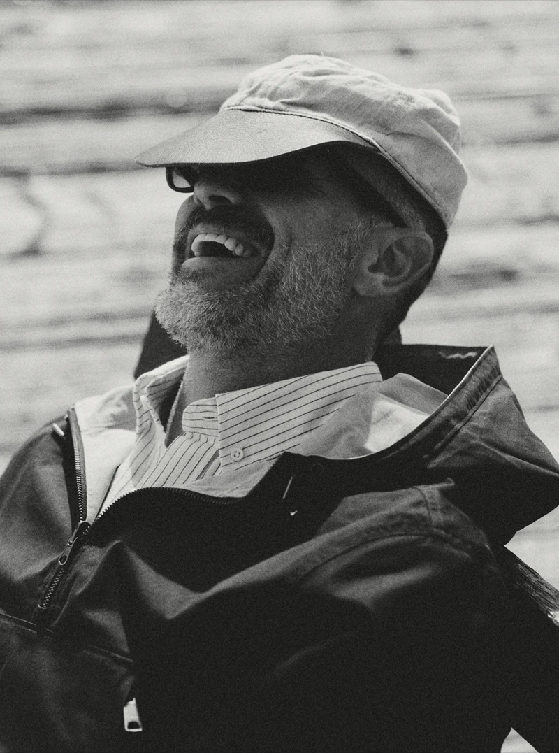 Black and white close-up of a man laughing in a cap and dark zip jacket layered over a striped shirt for Taylor Stitch summer 2026