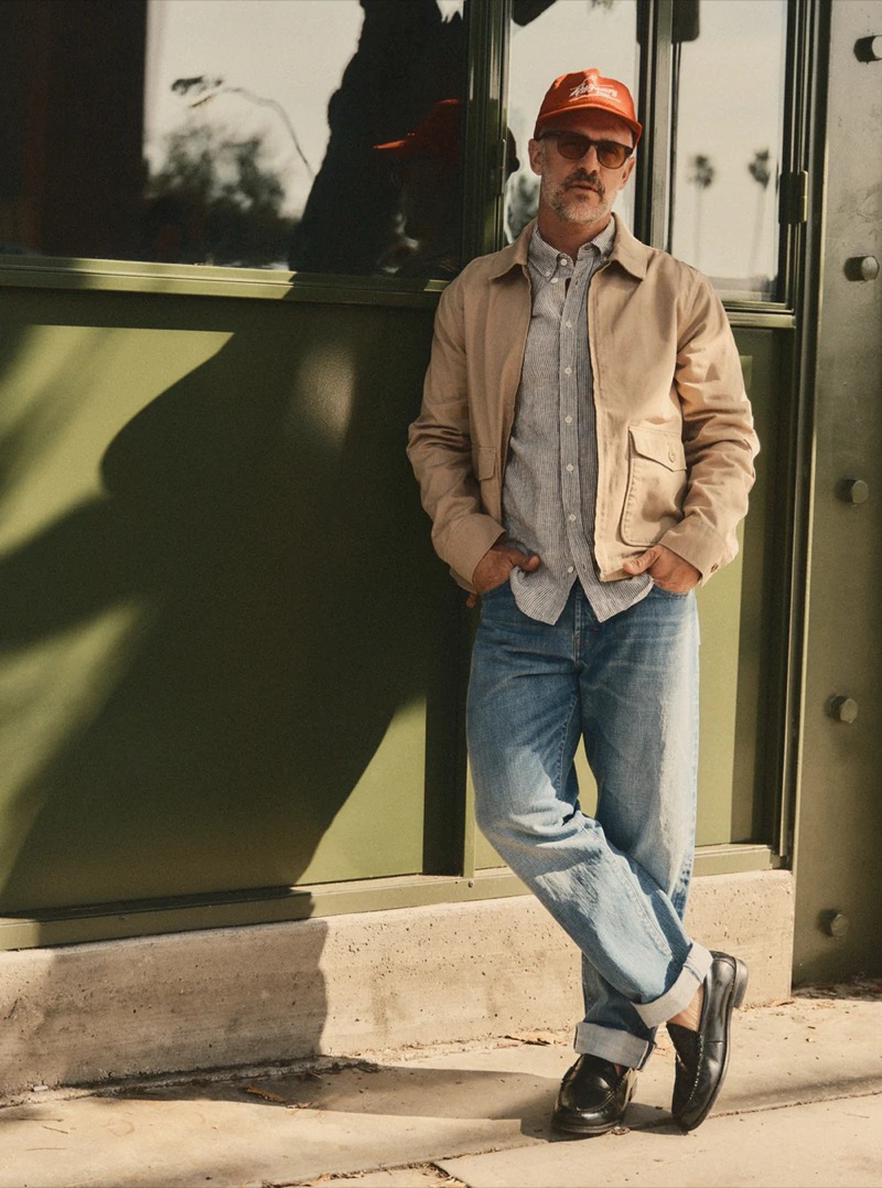 A man in a tan Harrington jacket over a gray chambray shirt, light wash cuffed jeans, and black loafers leans against a green storefront for Taylor Stitch summer 2026