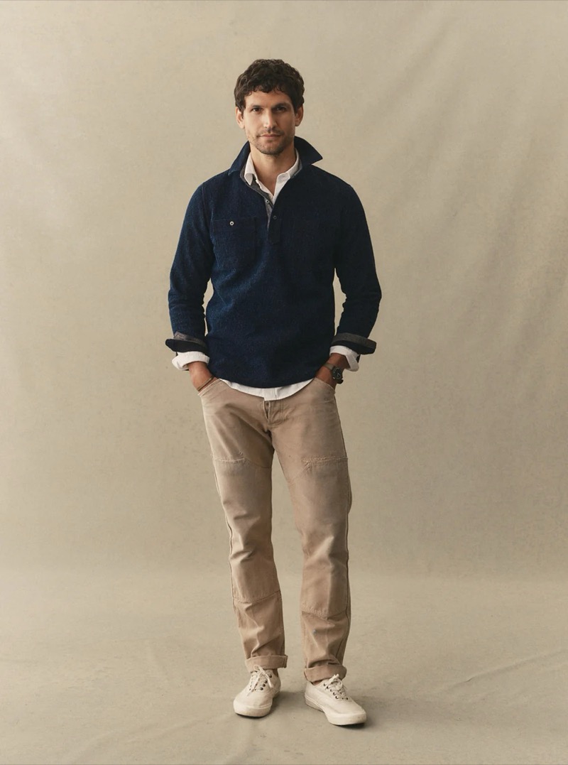 A man wears a navy snap-collar pullover layered over a white shirt with khaki utility trousers and white sneakers for Taylor Stitch summer 2026