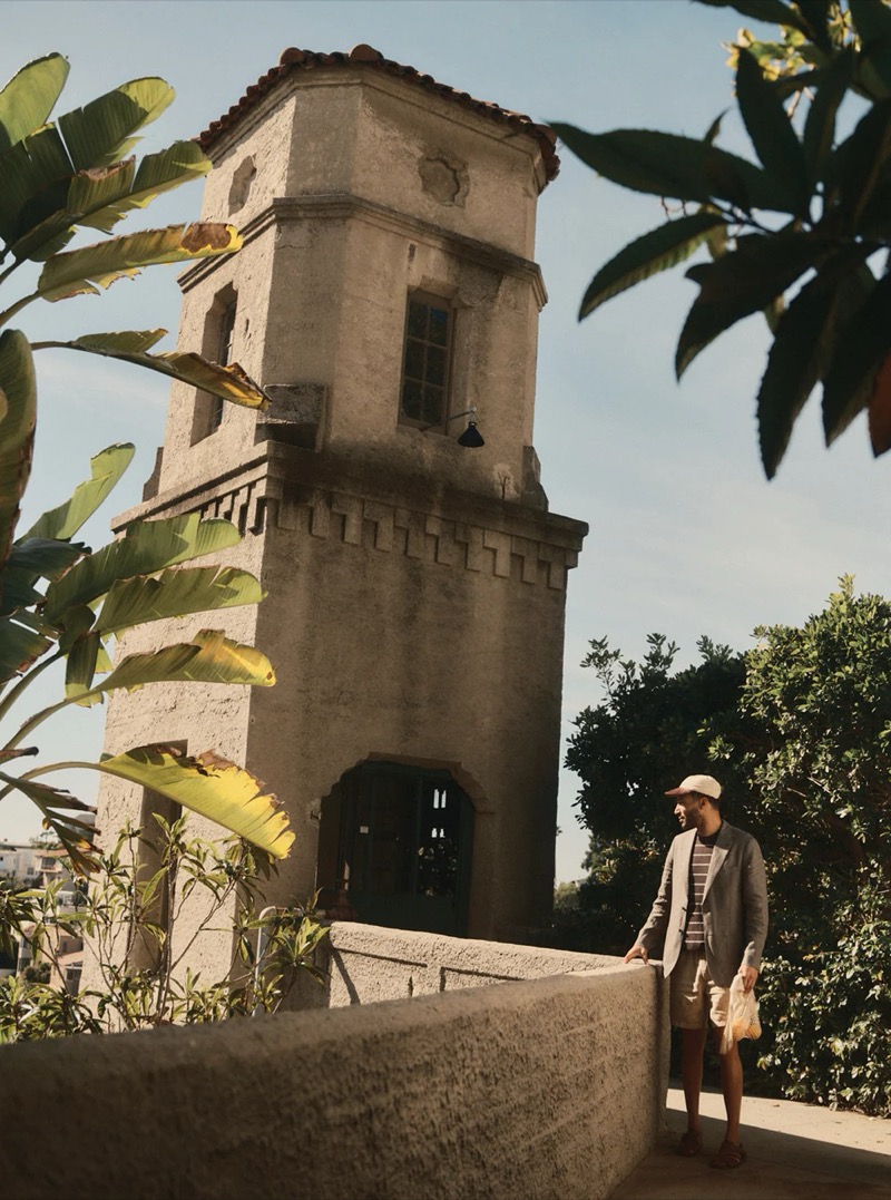 A man in a gray blazer over a striped t-shirt and linen shorts carries a net bag near a stone tower for Taylor Stitch summer 2026
