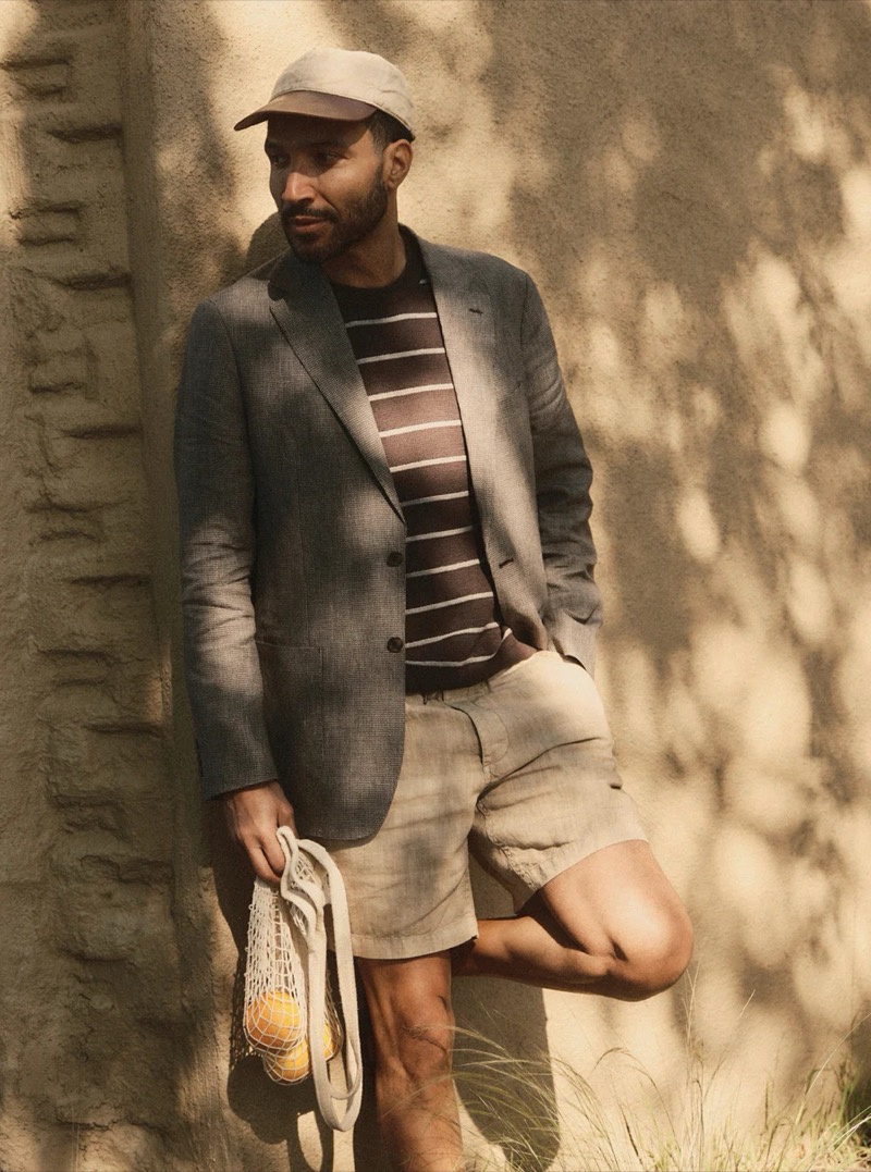 A man in a gray blazer over a striped t-shirt, linen shorts, and bucket hat leans against a sun-drenched stone wall for Taylor Stitch summer 2026