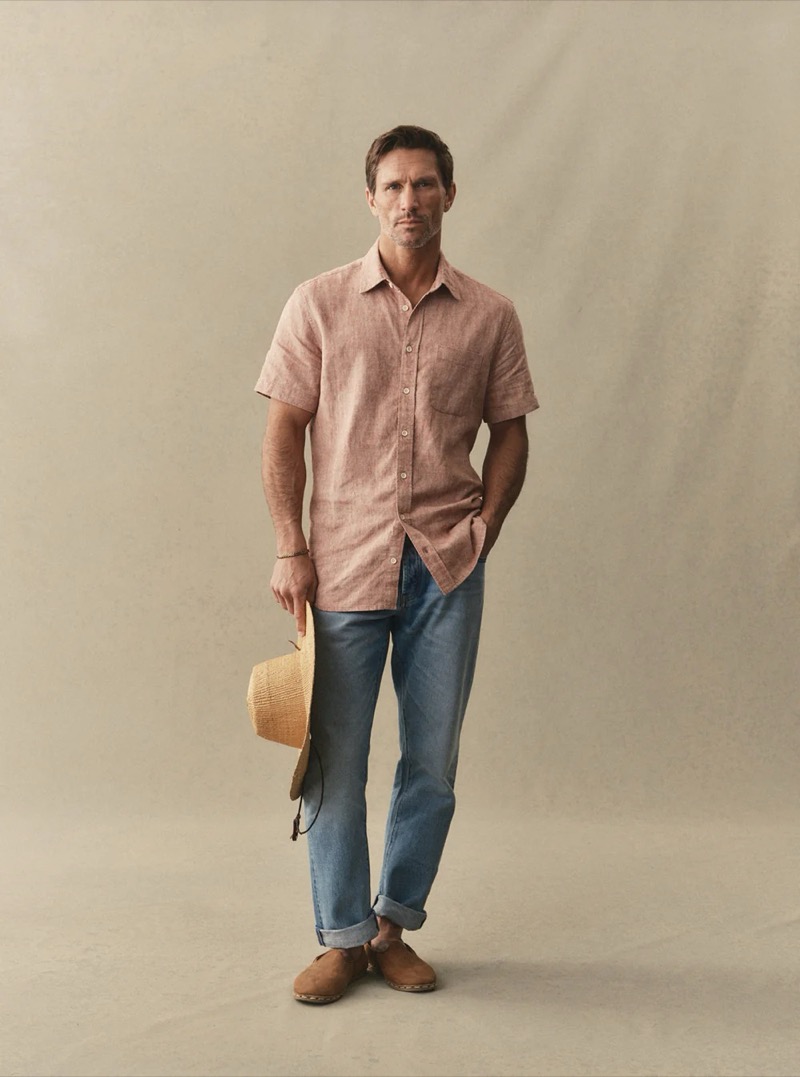 A man in a washed pink short-sleeve linen shirt, light wash cuffed jeans, and desert boots holds a straw hat for Taylor Stitch summer 2026