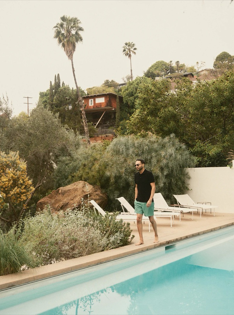 A man in a black polo shirt and green shorts stands by a pool surrounded by palm trees for Taylor Stitch summer 2026