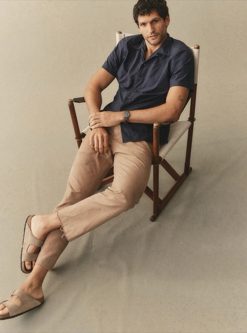 A man in a navy camp collar short-sleeve shirt and stone linen trousers sits in a canvas director's chair for Taylor Stitch summer 2026