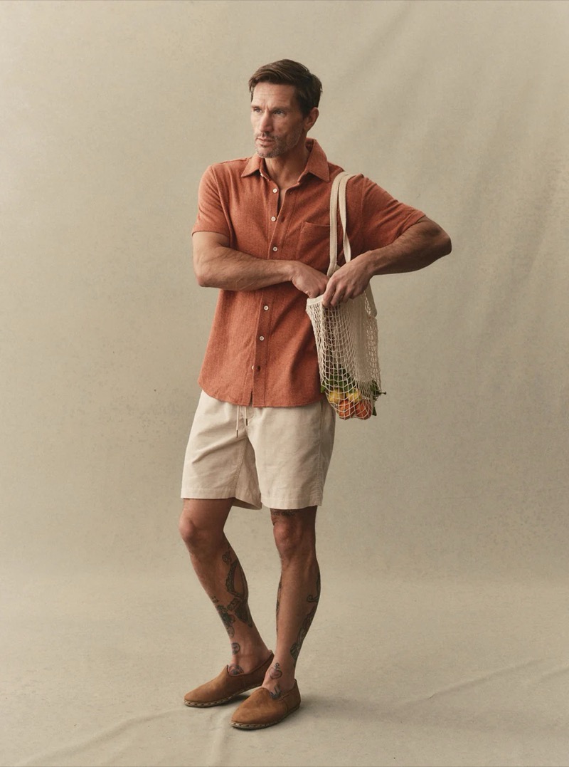 A man in a terracotta short-sleeve knit button shirt and cream drawstring shorts carries a net bag for Taylor Stitch summer 2026