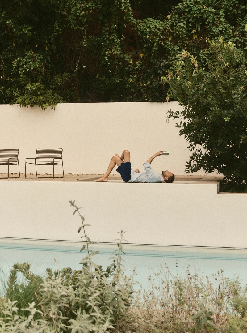 A man in a light blue shirt and navy shorts lies on a poolside terrace reading a book for Taylor Stitch summer 2026