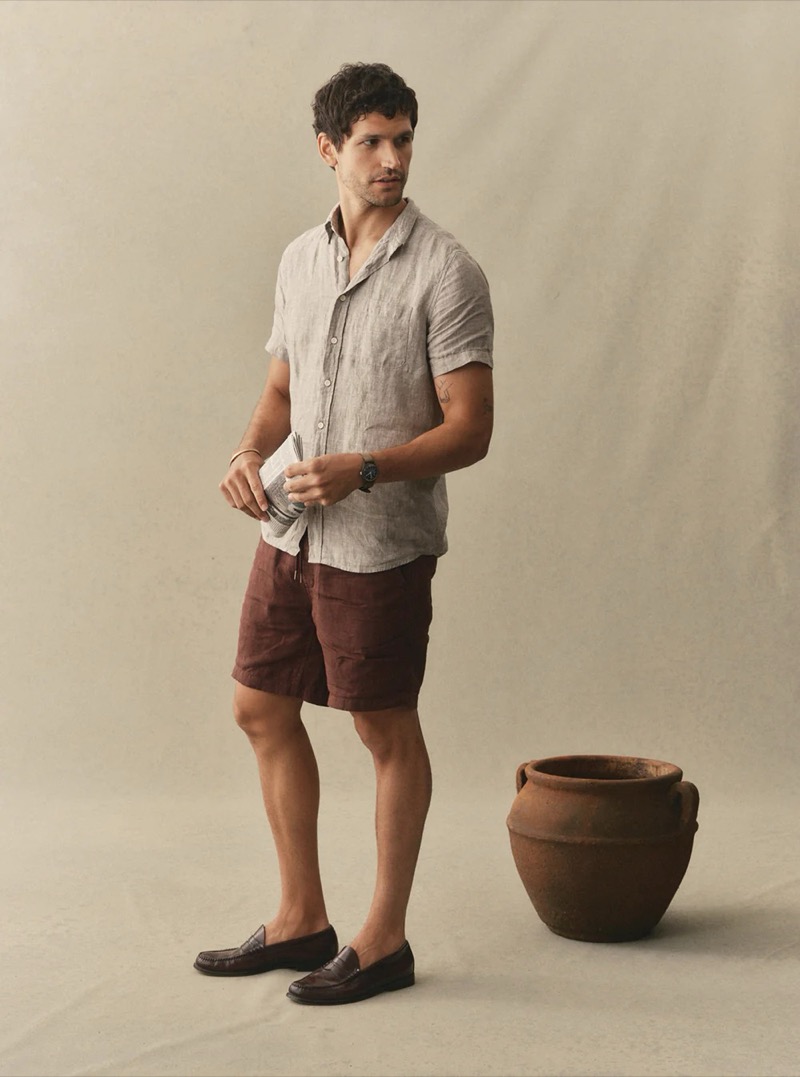 A man in a natural linen camp collar shirt and burgundy linen shorts with dark penny loafers stands beside a ceramic pot for Taylor Stitch summer 2026