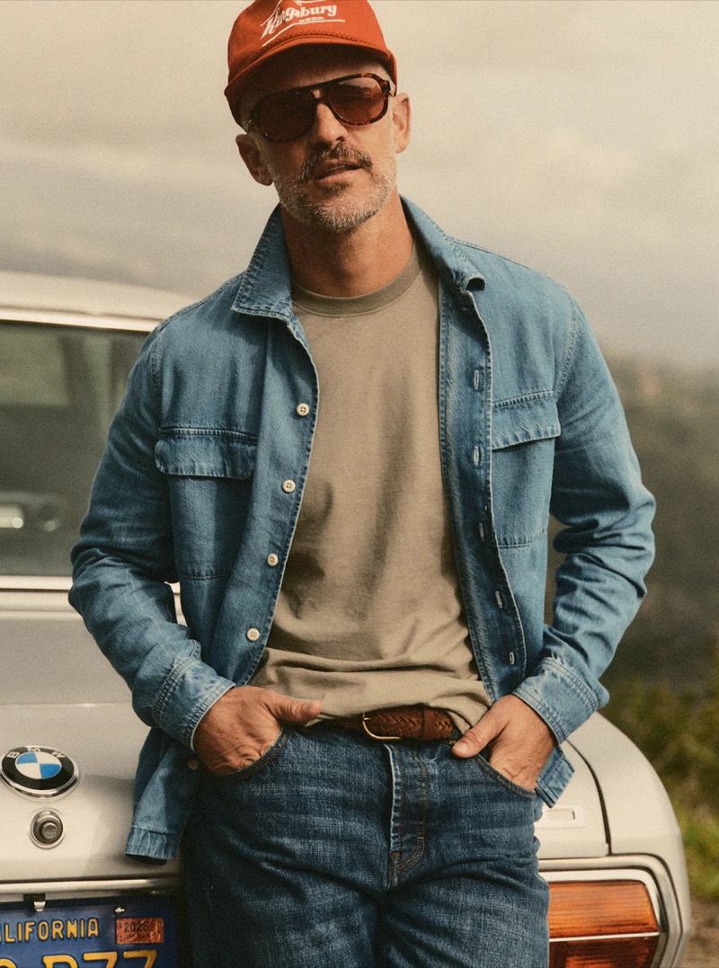 Close-up of a man in an open denim overshirt over a gray t-shirt with a red cap and sunglasses leaning on a vintage BMW for Taylor Stitch summer 2026