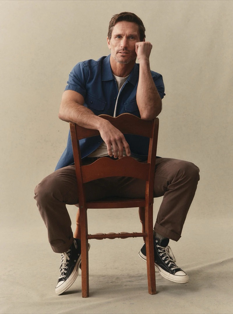 A man in a blue short-sleeve utility shirt and dark brown trousers sits astride a wooden chair in black high-top sneakers for Taylor Stitch summer 2026
