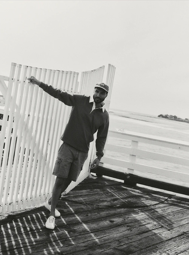 Black and white image of a man in a dark long-sleeve rugby polo, cargo shorts, and white sneakers leaning on a white fence on a seaside pier for Taylor Stitch summer 2026