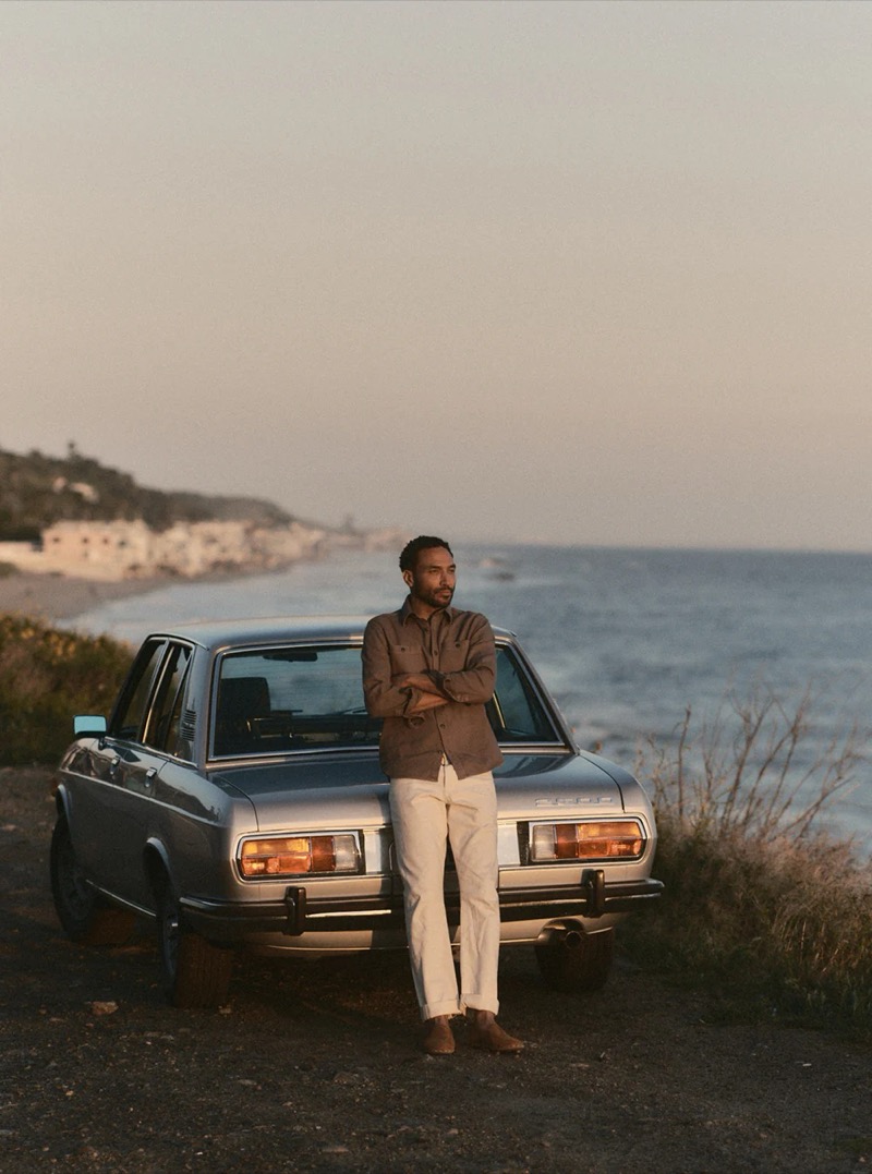A man in a brown utility overshirt and off-white trousers leans against a vintage silver sedan on a coastal road at dusk for Taylor Stitch summer 2026