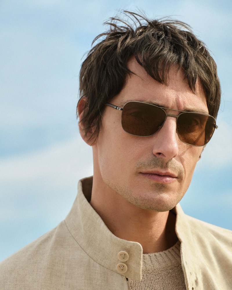 Davide wears gold aviator sunglasses with amber lenses and a sand linen band-collar jacket from Tod's against a bright summer sky